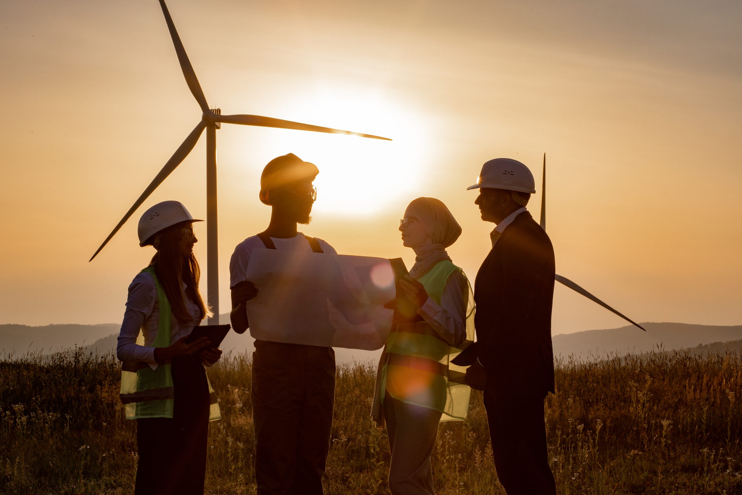 Multi ethnic group of engineers, technicians and managers in safety helmets having meeting outdoors about green energy. Men and women using tablet and drawing while working on windmill farm.